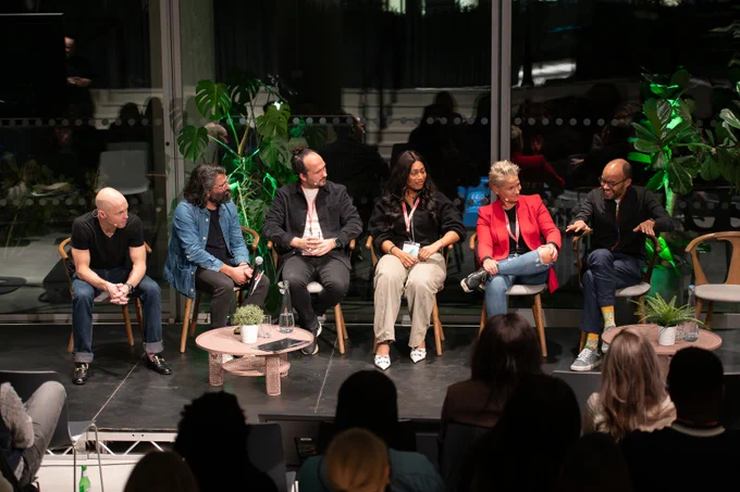 It shows six people in wooden chairs on a stage. On the far left is Dan a man in a black t-shirt, then it is Vinay in a denim jacket, then Nikki Wicks who is wearing a black jacket and jeans, then it is Grace who is wearing a black top and cream trousers, beside her is Sam who is wearing a pink blazer and has short blonde hair. They are all looking at Tom who is speaking and wearing jeans and a black jacket.