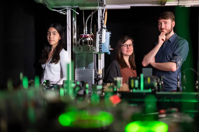 Full-color photo of P.I. Marissa Weichman and two lab members in the spectroscopy lab, with green lights in foreground. (Photo: Sameer Khan)