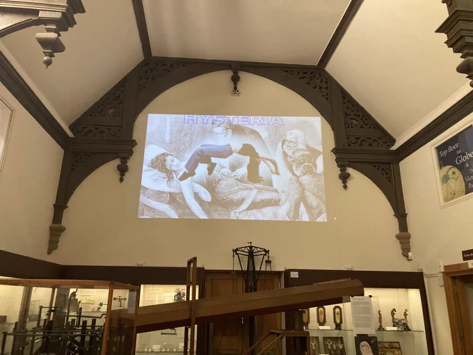 Projection on a wall above a telescope in a museum gallery showing a woman’s body superimposed onto a historic drawing of a woman lying down and the title ‘Hysteria’.