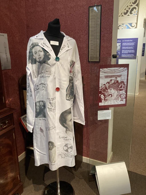On the left, mounted on a mannequin, a white lab coat with pictures of female scientists. On the right, mounted in a glass panel, a red and white print showing female scientists.