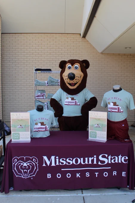 Boomer, the mascot standing behind the bookstore selling table in his 2024 Be A Bear shirt. 