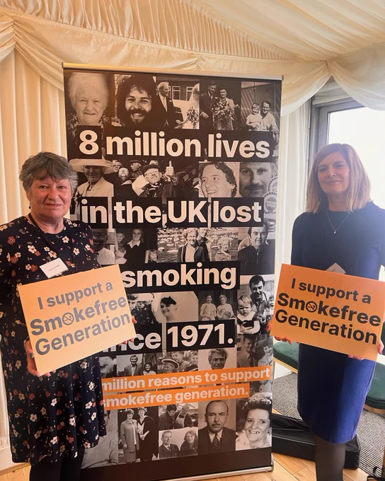 Liz (left) holding a sign which reads "I support a Smokefree Generation" and standing in front of a banner that says "8 million lives in the UK lost to smoking since 1971."