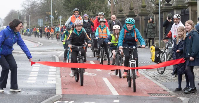 Children on bikes cycling along a segregated cycle lane towards the camera. A woman on the left and two girls on the right hold a ribbon in front of them