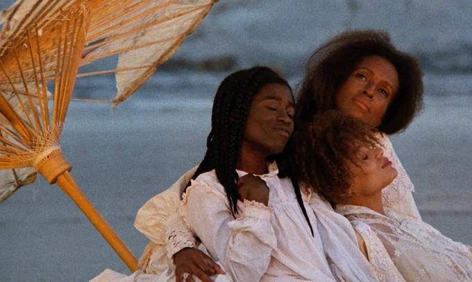 A still from the film Daughters of the Dust by Julie Dash shows three Black women of different ages leaning on each other, all dressed in white, under an umbrella, they look at peace. They seem to be near a body of water as well.