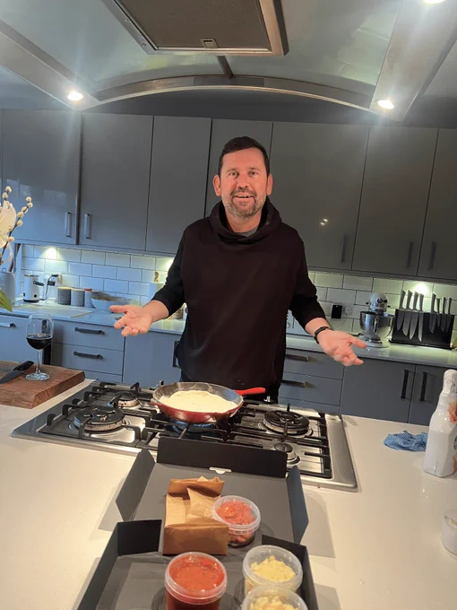 Man in black jumper stood in his kitchen - looking a bit confused! Pizza ingredients in the foreground.
