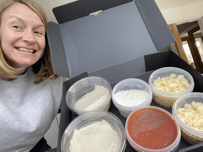 Woman in grey jumper smiling while holding up a box full of pizza ingredients 