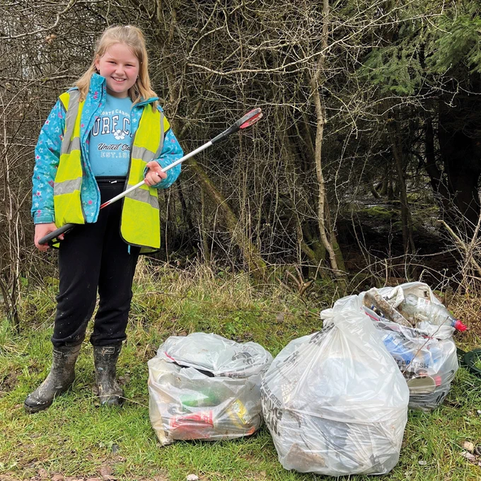 Photo of a young teenage litter picker. She is looking at the camera and smiling. She is holding a litter grabber and is stood beside three filled sacks of litter. She is stood on grass beside some trees and wears a hi-vis vest.