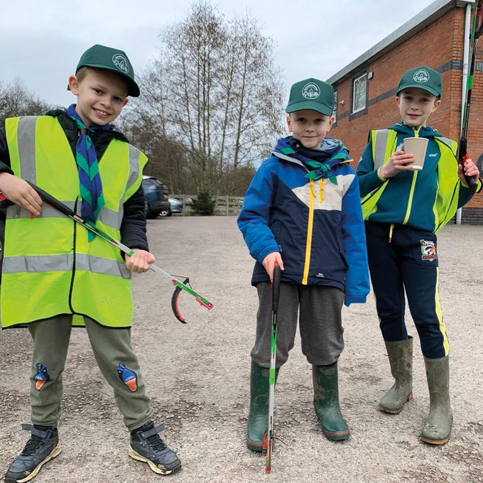 Photo of 3 boys in cub uniform. All 3 are wearing similar baseball style caps and holding litter pickers. They look happy.