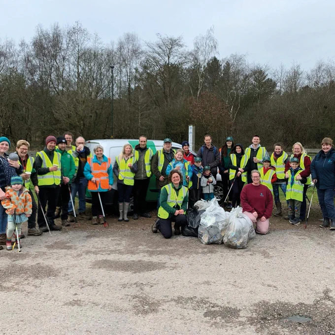 Group photo of many of the litter-picking volunteers. They are stood in a row around some of the bags of litter. Most are wearing hi-vis vests. They are smiling at the camera. In the distance is a row of trees.