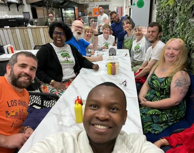 A group of 11 men and women sit inside a cafe around a long white tables. They wave and smile at a camera held by a young, smiling black man at the head of the table. Some are wearing CUE white t-shirts with the green tree logo on them. They all look happy.