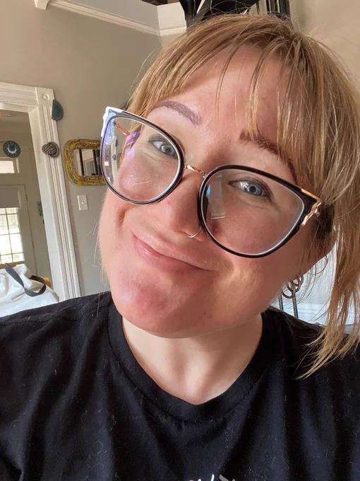 A blonde woman with black glasses smiles in a selfie. She has freshly microbladed eyebrows—you can see the brush strokes.