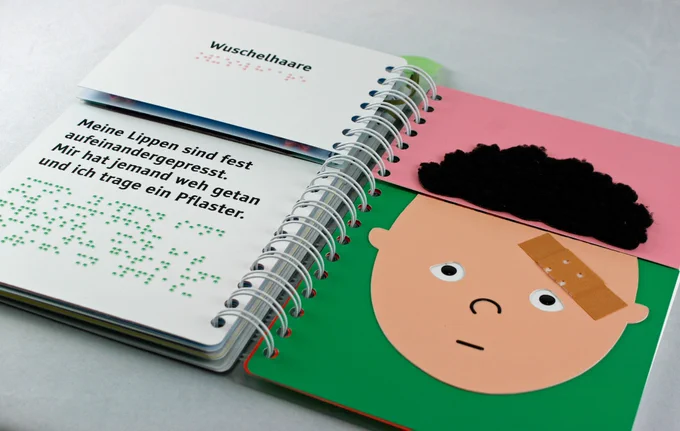 Aufgeschlagenes Ringbuch (rechts ein Gesicht mit Pflaster auf der Stirn und Wuschelhaaren). Links der Text in Großdruck und Braille.