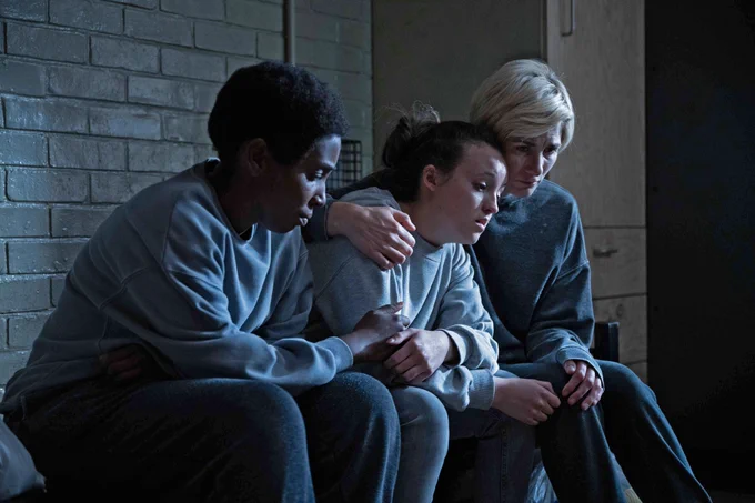 Image of Abi (Tamara Lawrance), Kelsey (Bella Ramsey) and Orla (Jodie Whittaker) they are sat on a bed in a prison cell embracing. 