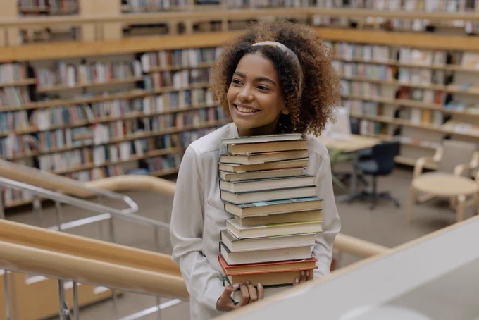 Young mixed race woman holding a pile of books in her arms in a library setting. 