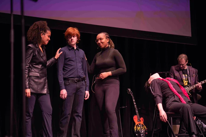 Performers converse next to an actor lying in a chair, with a musician in the background.