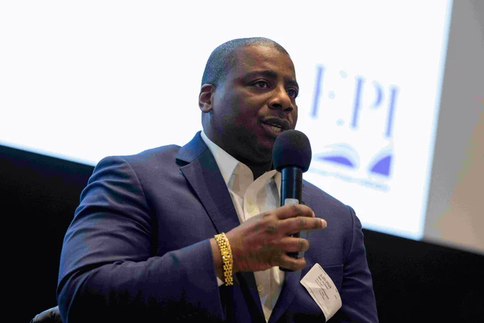 A person in a blue suit coat and white shirt speaks into a microphone.