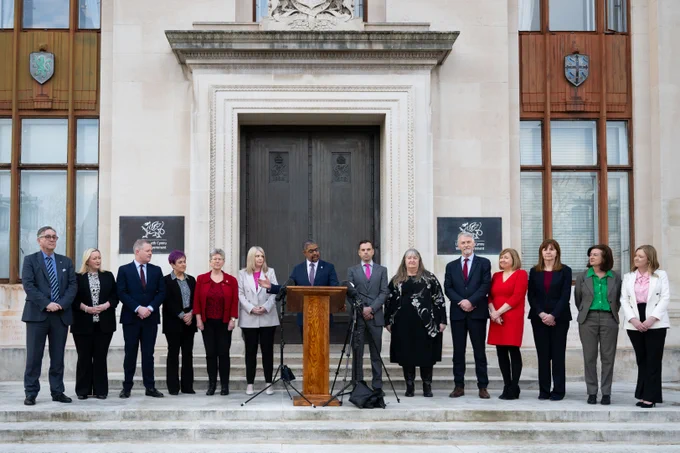 An image of the new welsh government cabinet. They stand together around the first minister outside the door of the Cathays Park offices