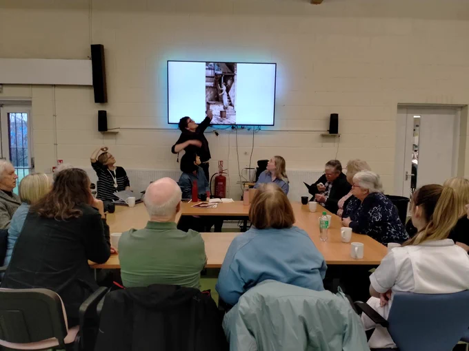 A group of people around a large table. They are looking a woman who is pointing to picture on a screen. The picture isn't clear.