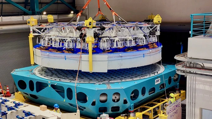 A thick white disk of glass with a blue protective coating on the top surface is held in the air by a complex web of white steel arms and suction cups. The glass is being lowered onto a teal steel platform with a large white circle in the middle.