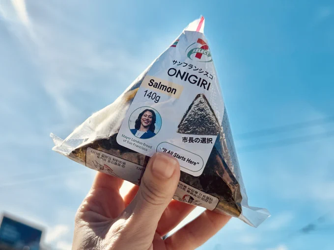 A person holding a packaged onigiri with a 7-Select label for salmon onigiri. On the label there's a picture of the mayor and next to it it says "It All Starts Here". There's also Japanese text that says サンフランシスコ (San Francisco) and 市長の選択 (Mayor's Choice).

