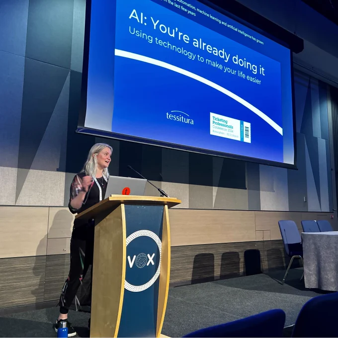 Photo of Ellie Griffiths at her 'AI: You're already doing it' session at the Ticketing Professionals Conference. Ellie is presenting at a podium with her laptop and looking at the crowd. There is a projected slide behind her that says 'AI: You're doing it already. Using technology to make your life easier.'