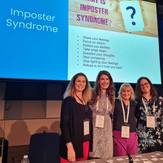 Photo of Rachael Easton, Kate Mroczkowski​, Lucy Costelloe and Sarah Bagg at their 'Expert NOT Imposter' session at the Ticketing Professionals Conference. The four women are standing side by side, smiling at the camera with a projected slide behind them that says: 'What is imposter syndrome?'