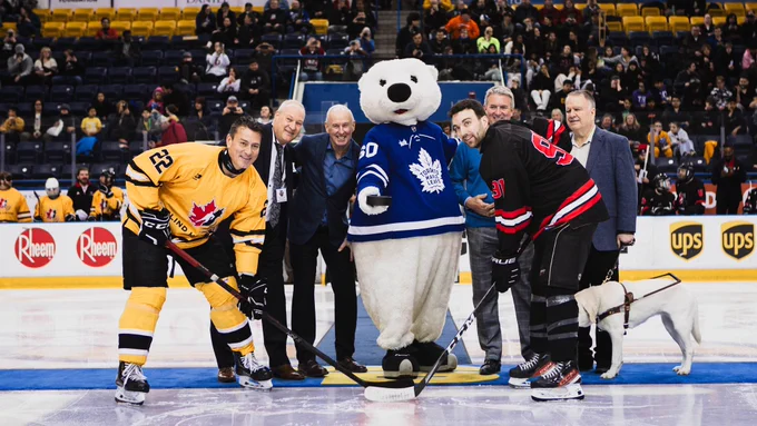 Puck drop with National Team captain Kelly Serbu and Jason Yuha, joined by Ron Maclean, John Rafferty, Bob Fenton and Carlton the Bear, hundreds of students are pictured behind them.
