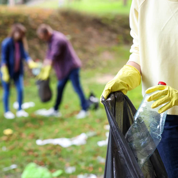 A close-up photograph of someone putting a plastic bottle in a bag. Two other people are also litter-picking in the background but they are blurred. 