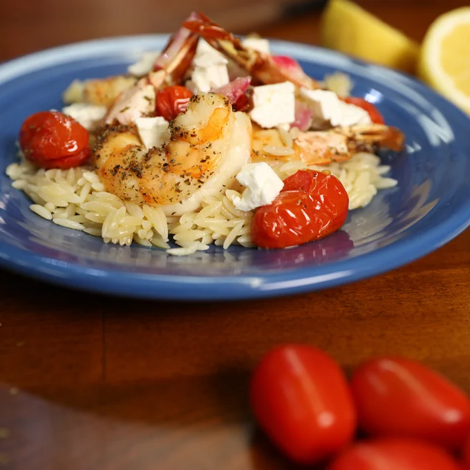 A blue dish of Shrimp With Orzo (Garides Me Kritharaki) sits on a table with tomatoes and lemon wedges