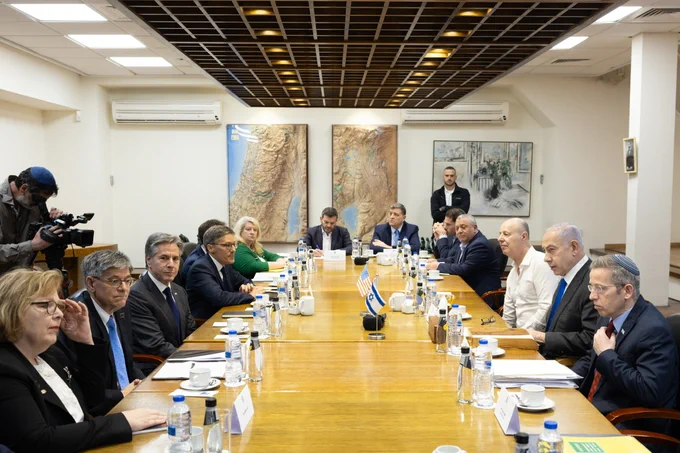 Secretary Blinken and U.S. officials sits across from Prime Minister Netanyahu and Israeli War Cabinet officials at a long wooden conference table lined with placards, water bottles, coffee cups, and note pads. 