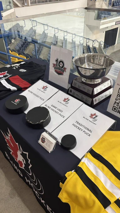A table display at the National Canadian Blind Hockey Tournament. On the left, an unused blind hockey puck is shown next to a game-used puck and a traditional hockey puck.