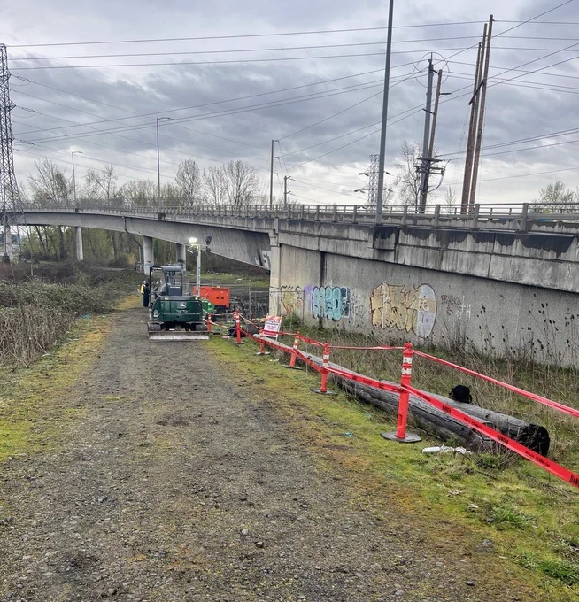 Red tape contains the site of a sewage release at the base of a concrete bridge overpass with graffiti. Heavy equipment in the background, soil and a gravel path in the foreground