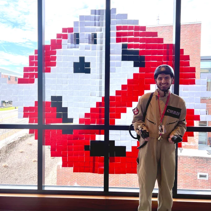 Chris is dressed in a Ghostbusters costume while standing in front of their logo made out of Post-it notes.