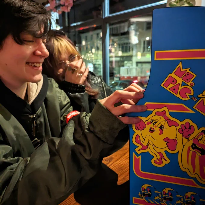 Two folks playing the Quarter Arcades Ms. Pac-Man. One is laughing a lot more than the other. A window in the background shows the blurred street outside.