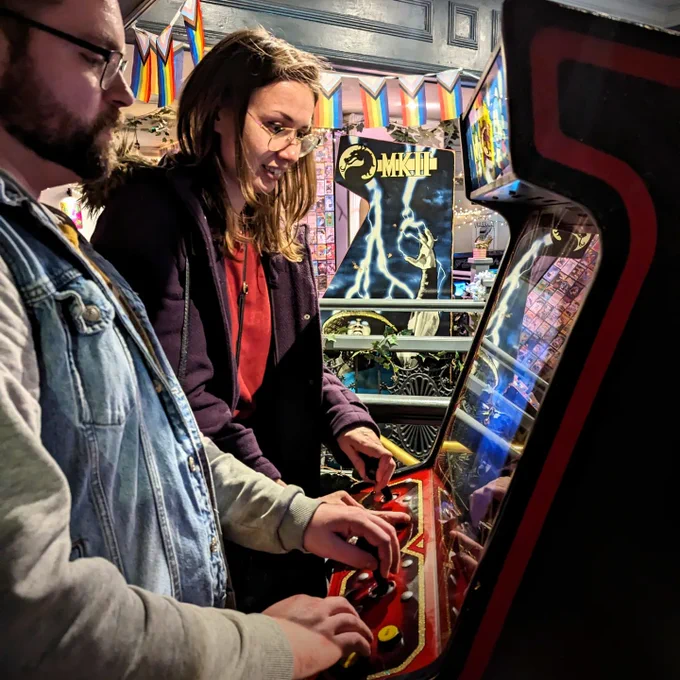 Two fine people playing Bubble Bobble. Mortal Kombat is visible in the background underneath some pride flags.