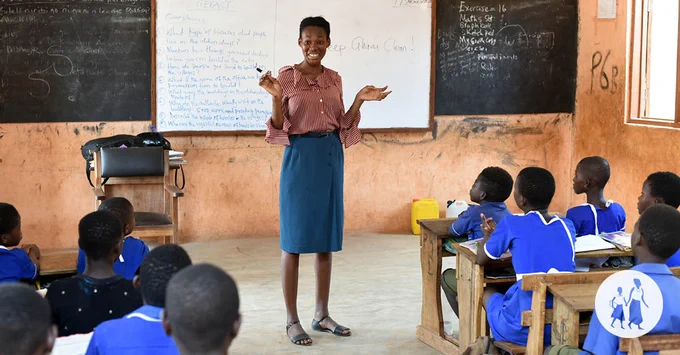  Cindy, a young Ghanaian woman, stands at the front of a classroom and speaks to a group of primary school students who are seated at desks and wear uniform.