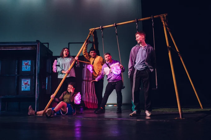 Five people are stood and sat around some swings. One individual is stood to the right and has a smug expression, the other four are looking at them, shocked. One of the individuals is sitting on the floor, with their hand up to their face and looking like they’re about to cry.