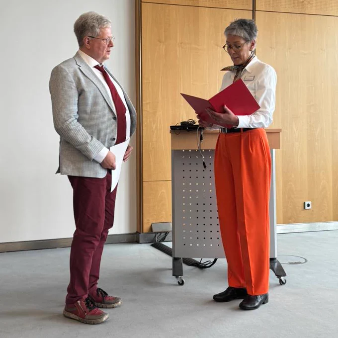 Prof. Dr. Thomas Kahlisch erhält die Urkunde von Karin Schmidt-Friderichs überreicht. Herr Kahlisch trägt eine rote Stoffhose, einen grau-karierten Blazer, ein weißes Hemd und eine rote Krawatte. Frau Schmidt-Friderichs trägt eine orangene Stoffhose und eine weiße Bluse, kurzes Haar mit Brille.