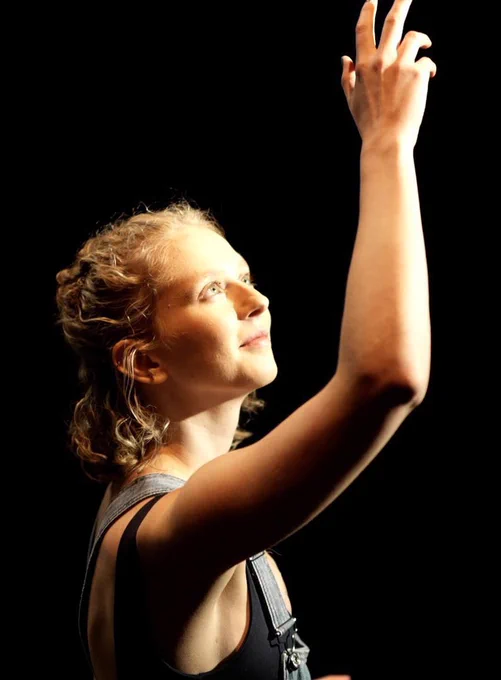 A young woman with blonde curly hair raises her arm towards a stage light. The light shines a warm glow over her face and body. She tilts her face towards the light and smiles.
