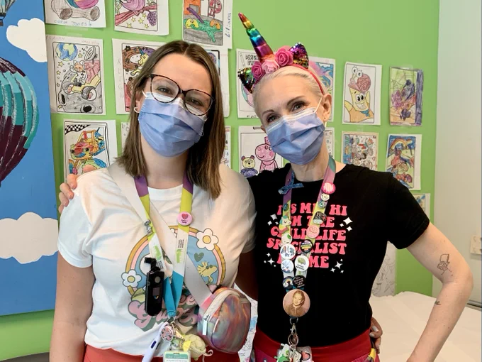 Two Child Life Specialists at Michael Garron Hospital wearing masks and standing in front of a colourful wall. 