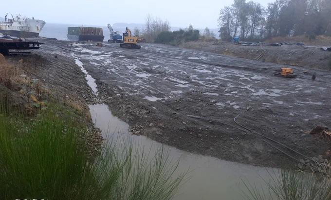 Pond in foreground with water running into it from muddy ground surrounding it. Ship, barge and industrial equipment in background.