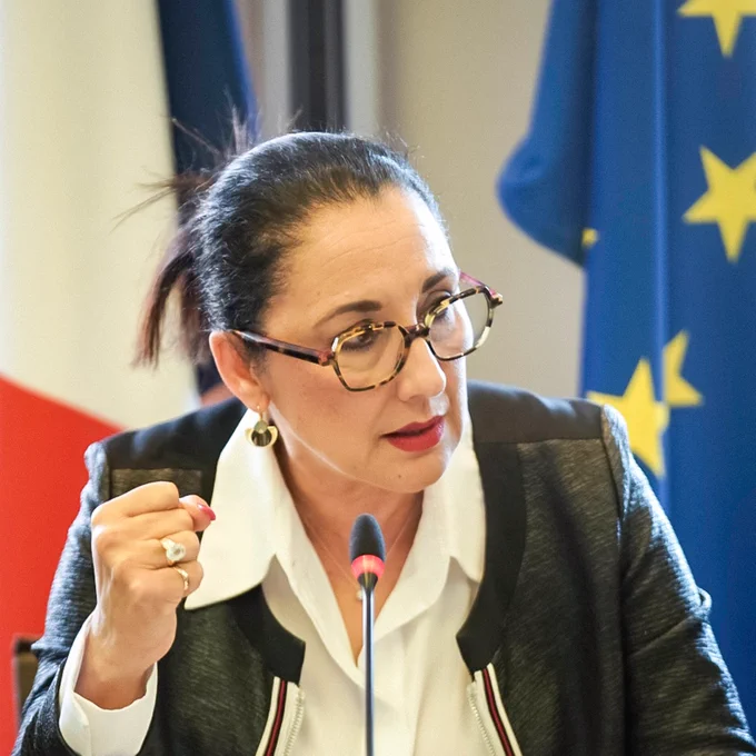 Photographie rapprochée de Fadila Khattabi. 

La ministre regarde à droite de l’image avec un point serré. Face à elle se trouve un micro, ainsi que des documents. Derrière elle se trouve le drapeau français et le drapeau européen.
