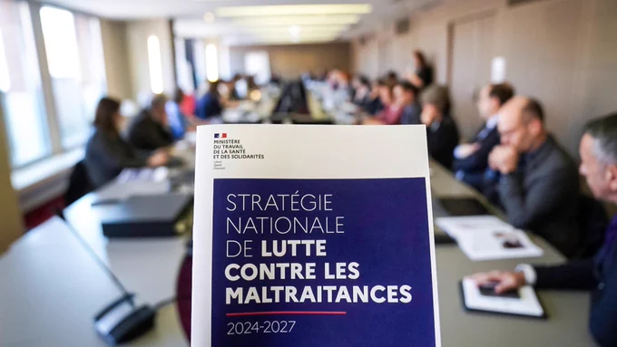 Au premier-plan se trouve le dossier de presse sur la stratégie nationale de lutte contre les maltraitances. 

L’arrière-plan est flouté. Les participants sont réunis autour d’une grande table de réunion. Des documents et des micros sont disposés sur celle-ci.