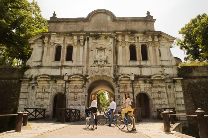Three bikers dressed in business casual clothes posing for the photo in front of a historical gate to the city of Parma, Italy.