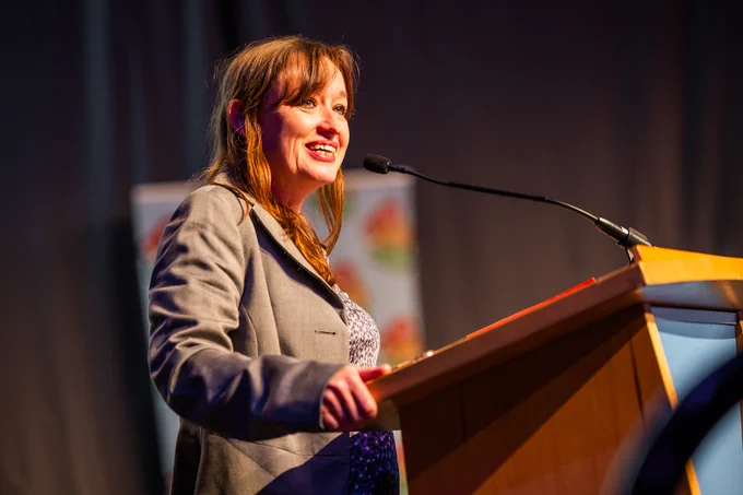 Emily Cherry is standing at a lectern at the Bikeability Conference 2024. She is smiling proudly at the audience.