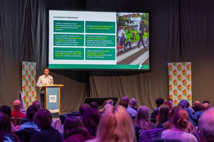 Guy Boulby from Active Travel England is presenting at the Bikeability Conference 2024. A slide showing active travel data is on a large projection screen.