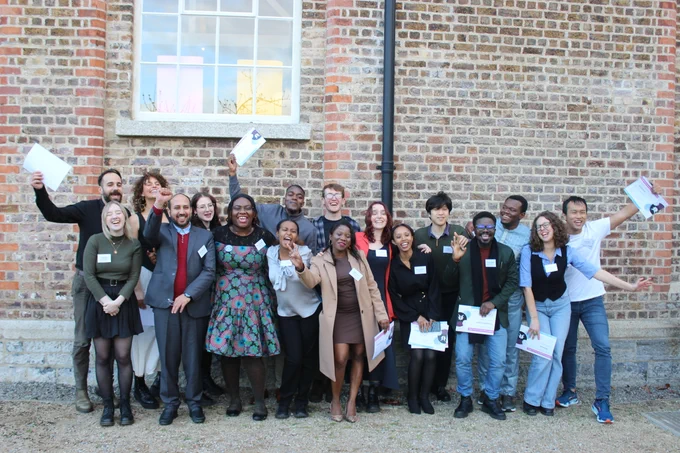 A photo of all the Changemakers with their certificates. There are about 20 of them, all different ages, races, and genders. They are standing next to a brick wall with a big window.