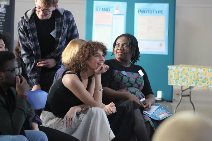 A candid photo of the audience at the Changemakers Showcase event. In the centre are Annie, a white woman with curly brown hair, and Wura, a black woman with short braids and a flowery dress. Annie is looking at the stage and smiling, her chin resting on her hand. Wura is looking away at someone out of shot. There are various other audience members in the background.