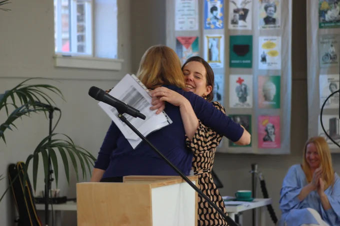 A photo of Clare and Nina, two white women, hugging. Clare is smiling, and Nina's back is to the camera. They are standing behind a lectern with a microphone on it. There are colourful posters and a window in the background.