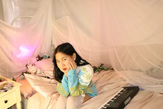 Promotional photo for "Super Fun Party Girl" which sees ÊMIA sitting on a bed with a keyboard out in front of her as she sits on the edge of the bed, looking at the camera with her dark hair over one shoulder, wearing a white-to-green-to-blue jumper and white stockings. There's a pink hue net over the bed and some small teddies on her pillow.